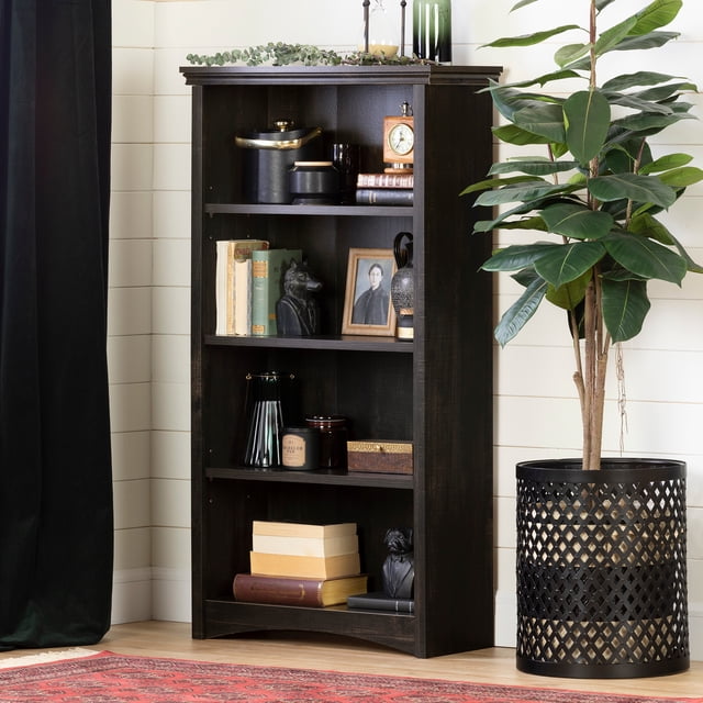 South Shore Gascony 4-Shelf Bookcase in rubbed black finish with four shelves, two adjustable, displaying books, decorative vases, candles, and framed photos, standing on a light hardwood floor next to a tall green plant in a black lattice planter against a white shiplap wall.