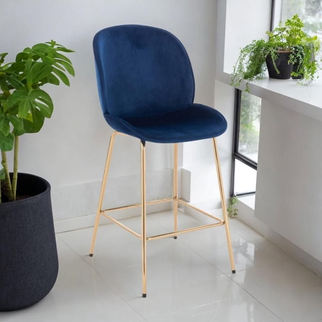 LOTUS GOLD STOOL (SLIM SEAT) with deep blue soft velvet upholstery and gold metal legs, featuring a slim curved seat and backrest, placed on white tile flooring near a window with green potted plants, ideal for kitchen or dining seating and often used as stylish vanity seating.