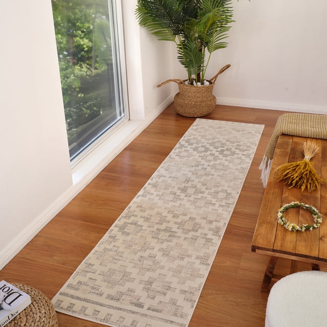 Rug Branch Astra Modern Indoor Area Rug in beige cream with an abstract Moroccan pattern, placed as a runner on a hardwood floor next to a large window. The 0.4-inch pile height rug features a jute backing for durability, suitable for high-traffic areas like hallways. Nearby are a woven basket with a green plant and a wooden bench with a yellow throw and floral wreath.
