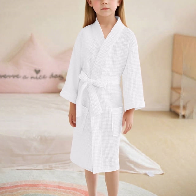 Kids' Long Sleeve Hooded Bath Towel Robe with Pocket, crafted from an absorbent polyester-spandex blend, displayed in a solid white color. This children's robe features long sleeves, a tie belt, and two front pockets, offering soft and breathable fabric ideal for bath, pool, and beach use. The child is standing in a cozy bedroom setting with soft furnishings in the background.