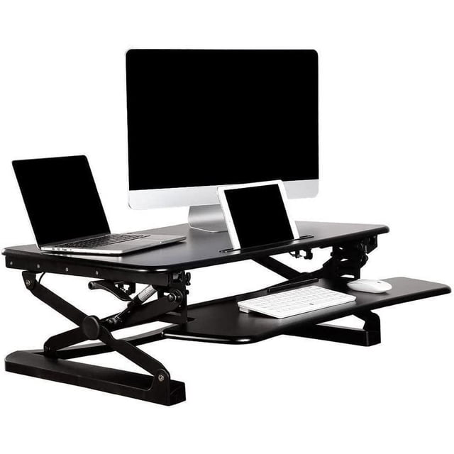 37 inch standing desk converter by PrimeCables, featuring a heavy-duty height-adjustable sit-stand desk riser with a keyboard tray. The black desk has a smart 'X' design with a gas spring mechanism, supporting up to 33 lbs and adjustable from 6 to 20 inches. The 35 x 23-inch platform accommodates dual monitors or a laptop and monitor setup, with a groove for smartphones or tablets. Includes a detachable keyboard tray and rubber pads to prevent scratches. Shown in an office setting with a desktop computer, laptop, and tablet.