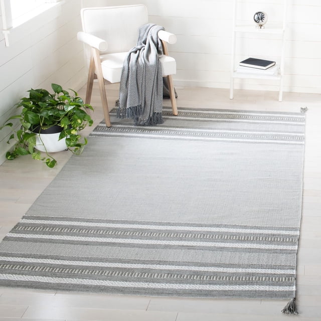 Safavieh Montauk Jobeth Striped Soft Braided Area Rug with flat-weave cotton texture in grey and white stripes and corner tassels, displayed on light hardwood flooring beside a white armchair with a grey throw and a green plant, suitable for coastal and contemporary interiors.