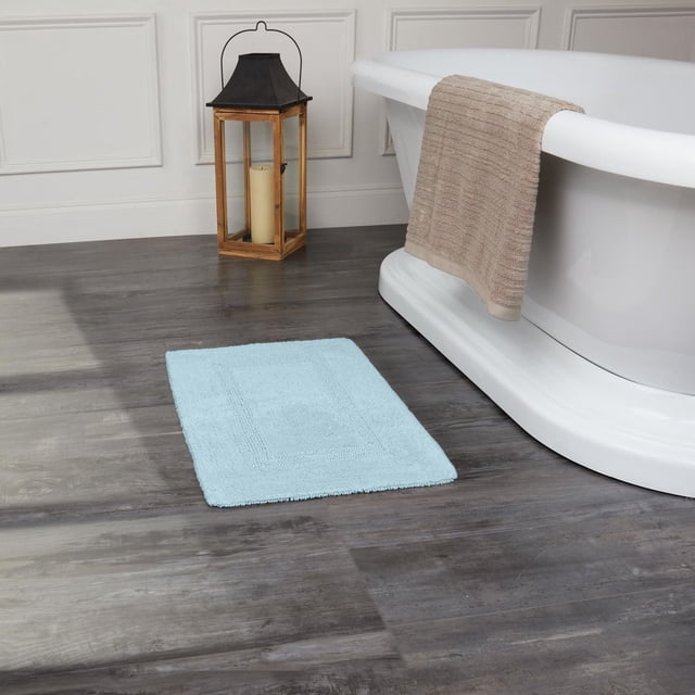 Fabstyles Basix Tapis de Bain en Coton Réversible Doux et Absorbant Bleu Pâle, Placé sur un Plancher de Bois Foncé, à Côté d'Une Baignoire Autoportante Blanche avec une Serviette beige Drapée sur le Bord et une Lanterne en Bois avec une Bougie Près d'Un Mur à Panneaux Blancs, Idéal pour la Douche Ou la Baignoire.
