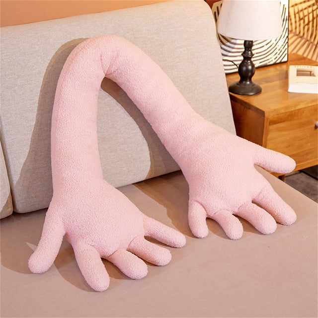 Funny Fleece Hand Pillow Cushion Toy Long Pillow Sleeping Leg Pillow in a soft pink color, featuring a unique design with two large hand-shaped ends connected by a long arm. Made of cotton, polyester, and velvet, this 31.5-inch pillow is ideal for hugging, snuggling, or as a playful decorative piece for imaginative-themed rooms. Displayed on a beige bed with a wooden nightstand in the background.