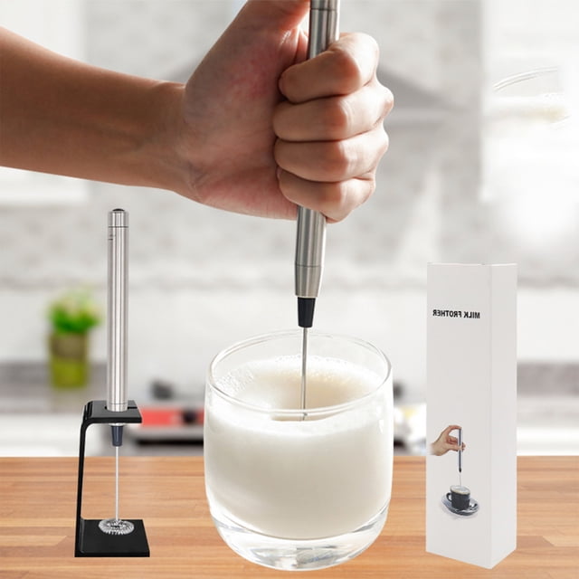 Dvkptbk Milk Frother Handheld, Battery Operated Electric Mixer in silver stainless steel with a sleek, compact design. The frother is shown in use, creating foam in a glass of milk, with its black stand and packaging visible on a wooden countertop. Suitable for lattes, cappuccinos, frappes, matcha, and hot chocolate, this handheld frother is a versatile kitchen tool.