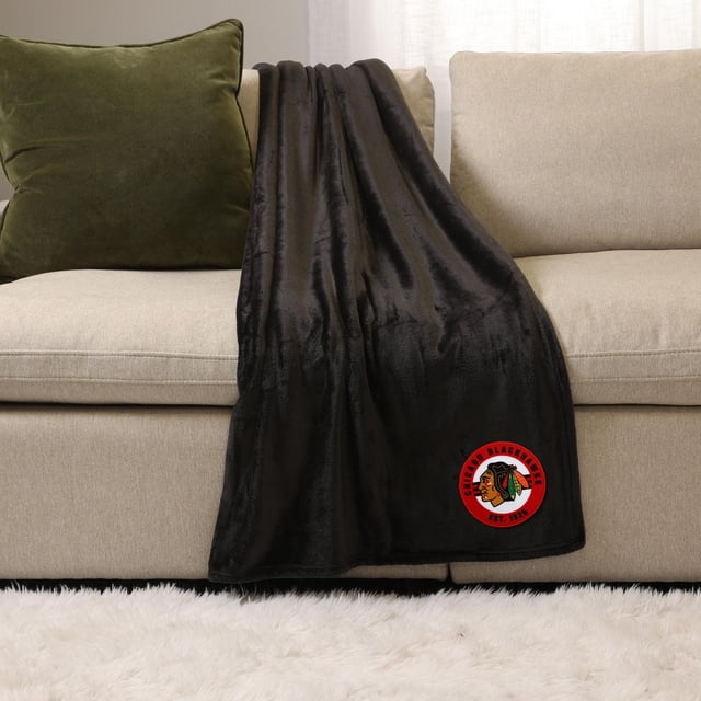 NHL, NBA, MLB Black Sports Throw Blanket, 50" x 60", draped over a beige sofa with a green pillow, made of soft plush 100% polyester fabric featuring a red Chicago Blackhawks logo patch, ideal for indoor use and cozy warmth.