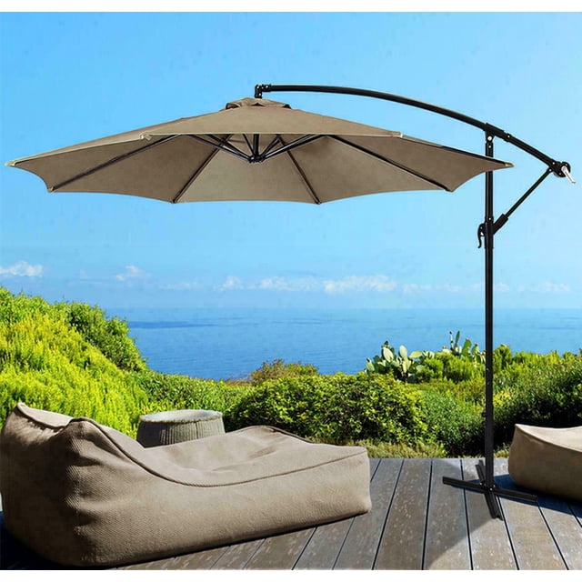 10Ft Offset Patio Umbrella with Cross Base and Crank, featuring a banana-shaped cantilever design and 8 sturdy ribs. The umbrella is made of UV-resistant, waterproof polyester fabric in a tan color, supported by a powder-coated steel pole with a 2.09-inch diameter. It includes a steel cross base for stability and a wind vent at the top for enhanced airflow. Shown in an outdoor patio setting overlooking a scenic ocean view, with lounge seating nearby.