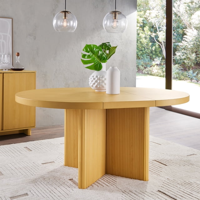 Better Homes & Gardens Lillian Fluted Round Expandable Dining Table in honey finish with a circular tabletop and fluted crossed base, expandable to 65.5 inches, set on a textured beige rug in a sunlit dining room featuring pendant lights and garden views, also suitable for versatile kitchen and dining spaces.