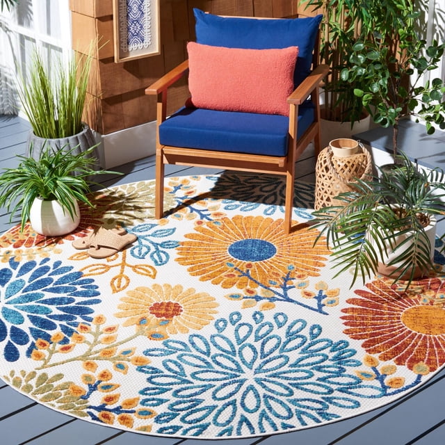 SAFAVIEH Cabana Tapis d'Extérieur en Grenat Floral sur une Terrasse Grise sous un Fauteuil en Bois avec des Coussins Bleus et un Oreiller Corail. le Tapis Rond Présente de Grands Motifs Floraux de Grenat, Moutarde et Bleu avec des accents Feuillus Verts. Il Est Entouré de Plantes en Pot et d'Une Lanterne Tressée, Convient aux patios Ou porches Extérieurs.