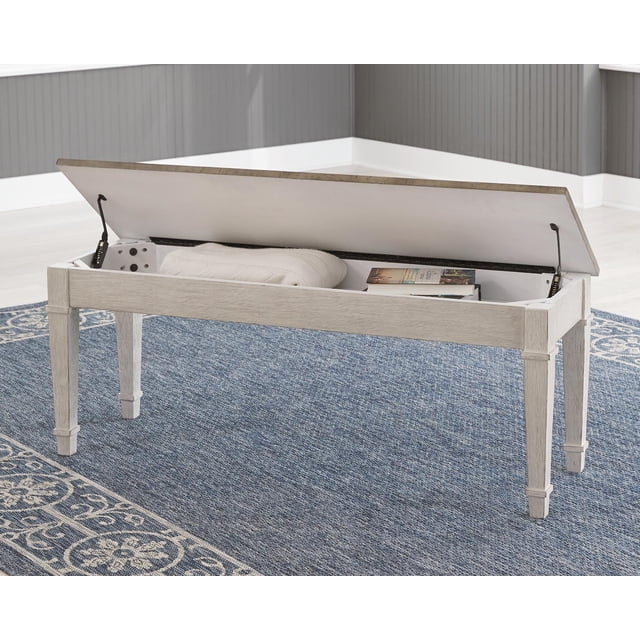 Signature Design by Ashley Skempton Banc de Rangement Style Ferme en Blanc et Brun Pâle, Conçu pour les Meubles d'Entrée. Présente un Dessus à Charnières, Espace de Rangement Intérieur Révélateur, Posé sur un Tapis à Motif Bleu dans un Décor d'Entrée Lumineux.