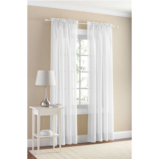 MAINSTAYS Marjorie Panneau de Rideau en Voile Uni en Tissu Voile Blanc Transparent Suspendu sur une Tringle Blanche au-Dessus d'Une Fenêtre avec des Carreaux à Grille dans une Pièce Murale beige, à Côté d'Une table d'Appoint Blanche avec une Lampe Argentée et un Bol Décoratif, et un Tapis Texturé Crème sur Plancher de Bois, Idéal pour Filtrer la Lumière et Adoucir les Couvre-Fenêtres, Souvent Utilisé pour Ajouter une Couche Délicate à la Déco de la Maison.