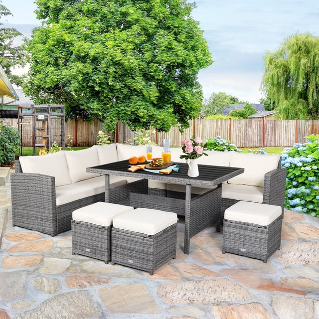 Patiojoy 7-Piece Wicker Patio Furniture Set featuring a mix grey rattan frame with off-white cushions. The set includes three corner chairs, three ottomans, and a rectangular dining table with a plastic wood top. The furniture is arranged on a stone patio in a backyard setting with lush greenery and flowers. Designed for outdoor use, the set offers a spacious and comfortable seating area, ideal for gatherings or casual conversations.