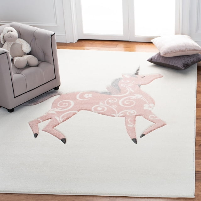 Safavieh Carousel Kids Dalach Unicorn Area Rug showing a pink unicorn with white floral swirl patterns on a white synthetic yarn background, placed on a hardwood floor beside a grey kids' armchair with a plush toy and two pillows, ideal for durable, soft kids' room decor.