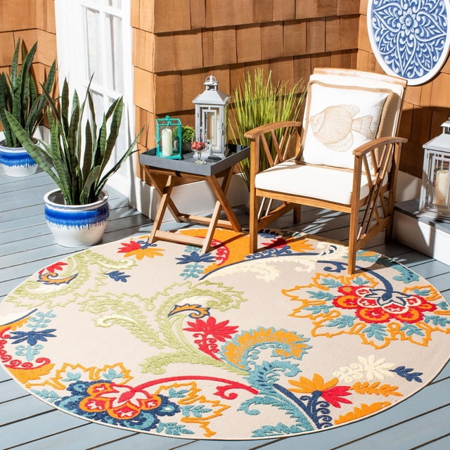 Safavieh Cabana Kuzman Floral Outdoor Area Rug, round with beige background and colorful floral and vine patterns in red, blue, green, yellow, and orange, placed on a gray wooden deck with a wooden armchair with white cushions and fish design pillow, a wooden side table with lanterns and glass bowl, surrounded by potted plants and brown shingle wall, ideal for patios or porches.