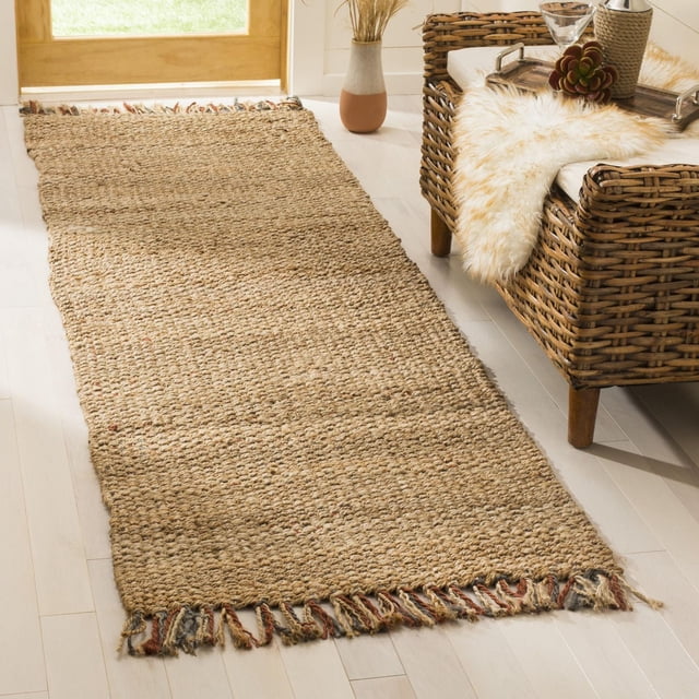 Safavieh Natural Fiber Gale Braided Area Rug in natural beige with multicolored braided fringe on both ends, laid on light hardwood floor beside a wicker armchair with white cushion and faux fur throw near a wooden door with sunlight, ideal for coastal or casual beach house style interiors.