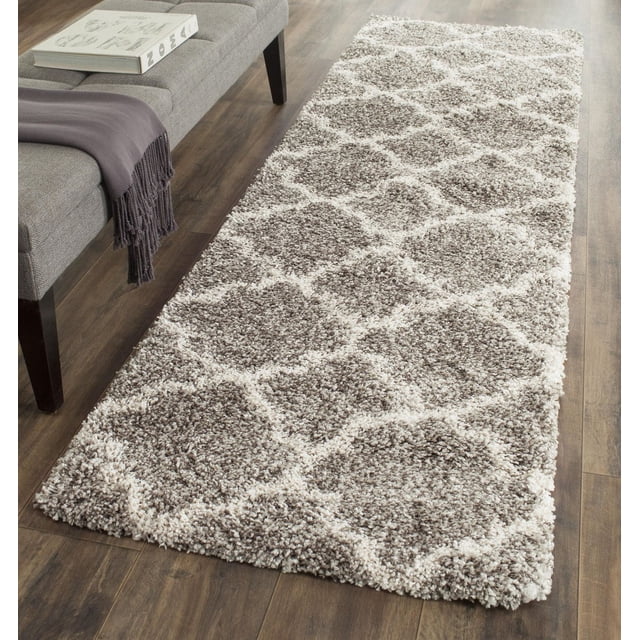 Safavieh Hudson Jaye Geometric Shag Area Rug on hardwood floor next to a grey upholstered bench with a dark grey throw and book, featuring a thick plush shag pile with grey and ivory onion-shaped tile patterns, suitable as an accent rug with durable, easy-care yarns for high traffic living spaces.