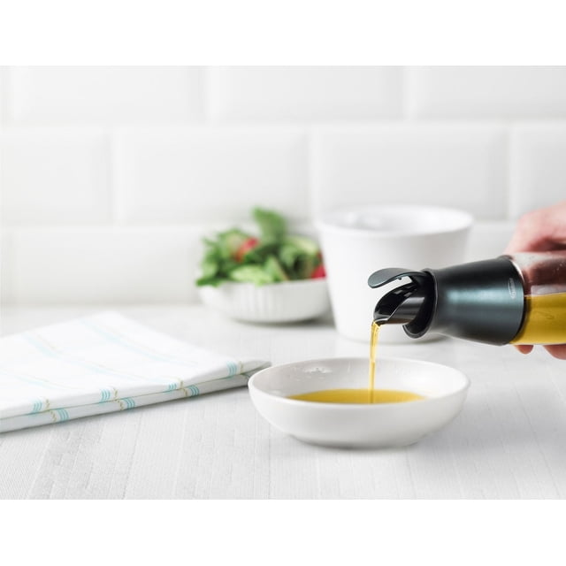 Trudeau Maison Automatic Oil Bottle with black dripless spout pouring oil into a white dish on a white kitchen counter, next to a folded white cloth and a bowl of salad in soft focus, ideal for precise oil or vinegar dispensing in food preparation.