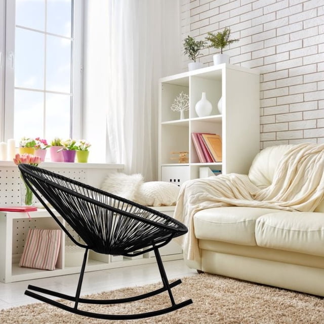 Costa Rocker Lounge Chair in black wire and plastic with oval seating, set in a bright living room with a cream leather sofa draped with a beige throw, white shelving with plants and books, and a large window with sheer curtains, suitable for indoor or outdoor use.