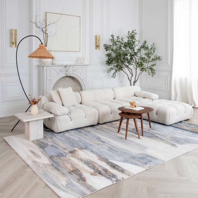 Rug Branch Atlas Mirage Indoor Area Rug in blue beige with abstract distressed pattern, displayed in a bright living room with a cream tufted sectional sofa, wooden nesting coffee tables, a marble side table with a vase, a curved rattan floor lamp, and a large green potted plant near a white decorative fireplace.