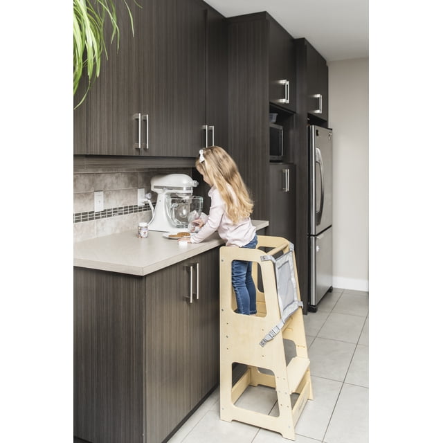 TREASURE KIDS Wooden Learning Tower for Toddlers 2 in 1, shown in a kitchen setting with a child standing on the natural wood step stool. The tower features a height-adjustable platform, a removable safety net, and a built-in whiteboard. Designed for Montessori-style learning, it allows children to safely participate in activities like cooking and crafts at counter height. The modern design integrates seamlessly into home spaces.
