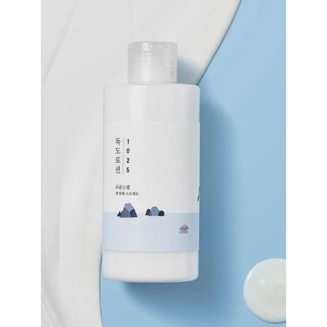 Round Lab 1025 Dokdo Lotion in a white cylindrical bottle with a clear flip-top cap, featuring a minimalist label with blue and gray rock illustrations on a light blue background, placed on a light blue surface with a white lotion smear and dollop, a moisturizing body lotion designed to balance skin hydration and soothe with natural minerals.