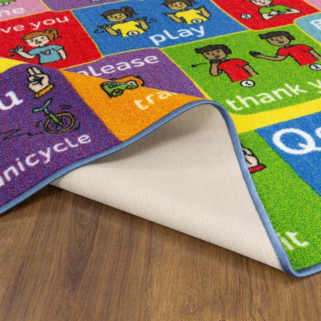 KC Cubs Playtime Collection ABC Alphabet ASL Sign Language Educational Learning & Game Area Rug Carpet for Kids and Children Bedrooms and Playroom showing colorful squares with American Sign Language hand signs and words like 'play,' 'thank you,' and 'unicycle,' with a folded corner revealing a beige non-slip backing on a wooden floor, suitable for educational play spaces.
