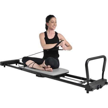 Stamina AeroPilates 287 Folding Pilates Workout Reformer Machine without Rebounder, featuring a wide cushioned platform, high-density foam shoulder pads, and adjustable headrest. A woman demonstrates a seated Pilates exercise using the machine's resistance cords. The reformer is black with a compact, foldable design and includes wheels for easy storage and transport. Ideal for upper body, lower body, and core workouts with a weight capacity of up to 300 pounds.