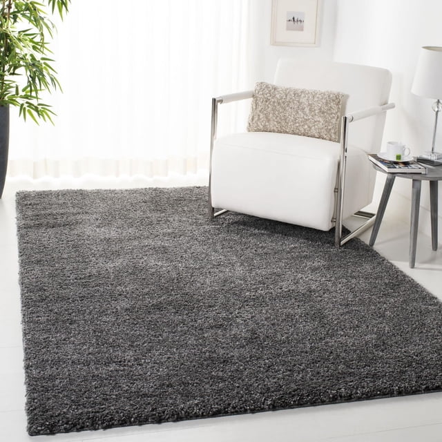 SAFAVIEH August Carlene Solid Plush Shag Area Rug in grey with a thick polypropylene shag pile, displayed on a white floor in a sunlit room. The rug complements a white armchair with chrome arms and a beige textured pillow, beside a round side table with a lamp and cup, reflecting a casual West Coast style. This plush accent rug is also suitable for cozy reading nooks or lounge areas.