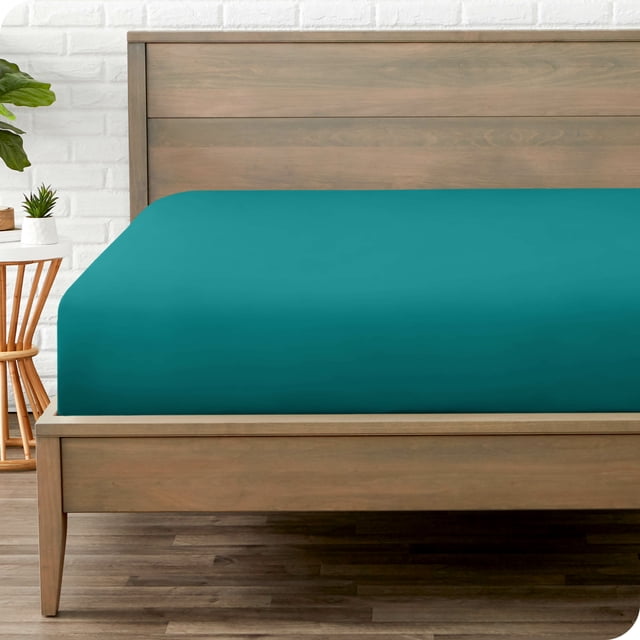 Bare Home Fitted Sheet from the Premium 1800 Collection in Twin XL size, shown in emerald green. The ultra-soft, wrinkle-resistant microfiber sheet is displayed on a wooden bed frame in a minimalist bedroom setting. Designed with deep pockets to fit mattresses up to 15 inches deep, this sheet offers a secure fit and hypoallergenic properties. Ideal for year-round comfort, it is machine washable for easy care.