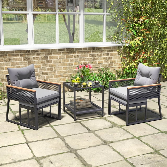 Costway 5 Piece Outdoor Conversation Set Rattan Armchairs with 2-Layer Side Table. This patio furniture set features two black PE rattan armchairs with gray cushions, a double-layer coffee table with a tempered glass top, and two ottomans that fit under the chairs for space-saving. The acacia wood armrests and reinforced metal frame add durability and style. The set is shown on a stone patio with a brick wall and lush greenery in the background.