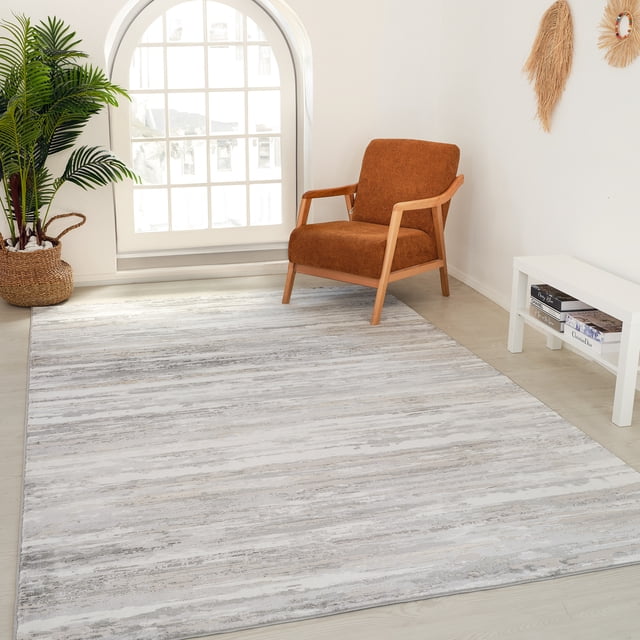 Rug Branch Atlas Lines Indoor Area Rug in beige grey with abstract horizontal stripe pattern on light wood floor, displayed in a bright room with a rust-coloured wooden armchair, large arched window with white grid panes, woven basket planter with green palm, and white console table with books underneath.