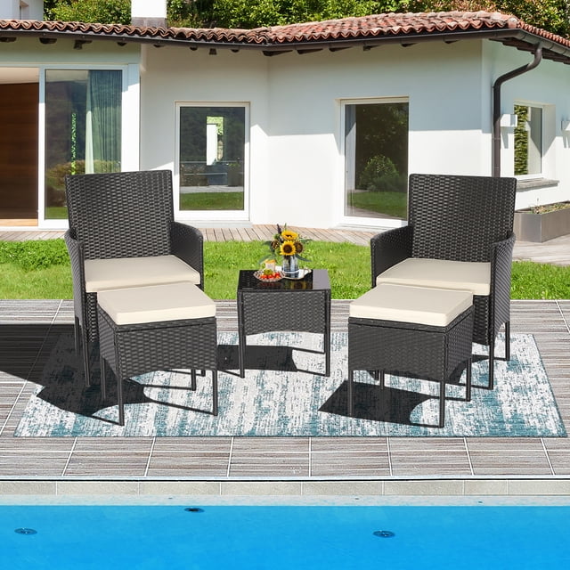 Costway 5PCS Patio Rattan Furniture Set Cushioned Sofa Ottoman Armrest Side Table Garden. This 5-piece set includes two black rattan single chairs with off-white cushions, two matching ottomans, and a black tempered glass coffee table. The ergonomic chairs feature curved armrests and cozy backrests, while the ottomans can double as footrests or additional seating. The set is displayed outdoors on a patio near a pool, with a modern house in the background.