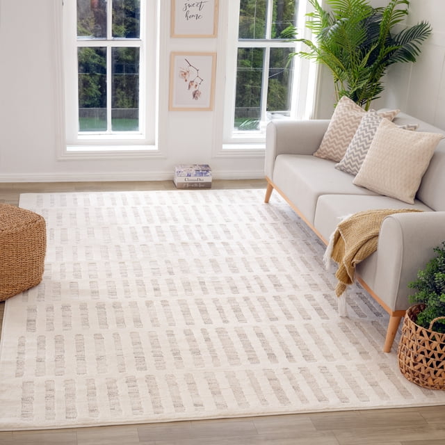 Rug Branch Astra Modern Indoor Area Rug in beige cream with an abstract pattern, laid on light wood flooring in a bright living room featuring a gray sofa with patterned cushions, a woven ottoman, green plants, and framed wall art between two windows, ideal for high-traffic areas like kitchens and hallways.