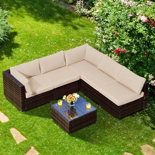 Costway 6PCS Patio Rattan Furniture Set Cushioned Sofa Coffee Table Garden Brown. The set includes 2 armless sofas, 2 corner sofas, and 1 coffee table with a tempered glass top. Made of durable PE rattan and a sturdy steel frame, it features beige cushions with removable covers for easy cleaning. The modular design allows for customizable arrangements, ideal for patios, gardens, or balconies. Shown in a lush green outdoor setting with flowers and grass.