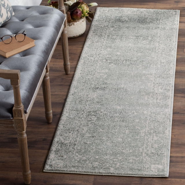 Safavieh Evoke Denica Traditional Area Rug with a muted grey and off-white distressed pattern and soft frieze texture, placed on dark hardwood floor next to a tufted grey bench with wooden legs and a book with glasses, suitable as an accent rug for transitional home decor.