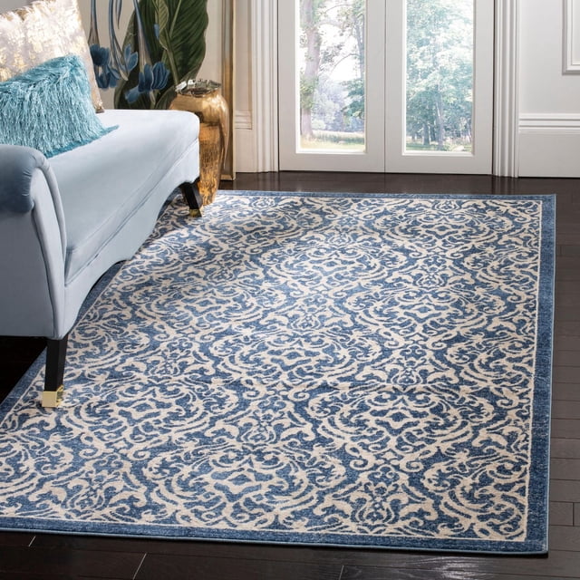 SAFAVIEH Brentwood Dilara Damask Area Rug with a close cut synthetic yarn pile in blue and cream damask pattern, placed on dark hardwood flooring beside a light blue sofa with decorative pillows in a bright room with glass doors, ideal for traditional and transitional decor and often used as an accent rug to add warmth.