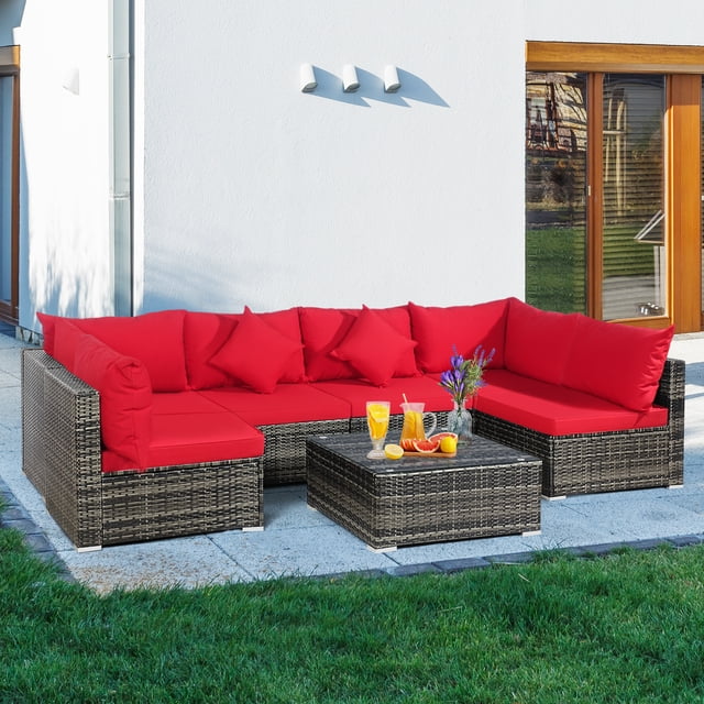 Patiojoy 7 Pcs Outdoor Patio Furniture Set featuring a mix gray PE rattan frame with vibrant red cushions. Includes a modular design with 2 corner sofas, 4 single sofas, and a coffee table with tempered glass. The set is arranged on a patio with a grassy lawn in the foreground and a modern white house in the background. Designed for outdoor use with weather-resistant materials and rustproof aluminum feet.