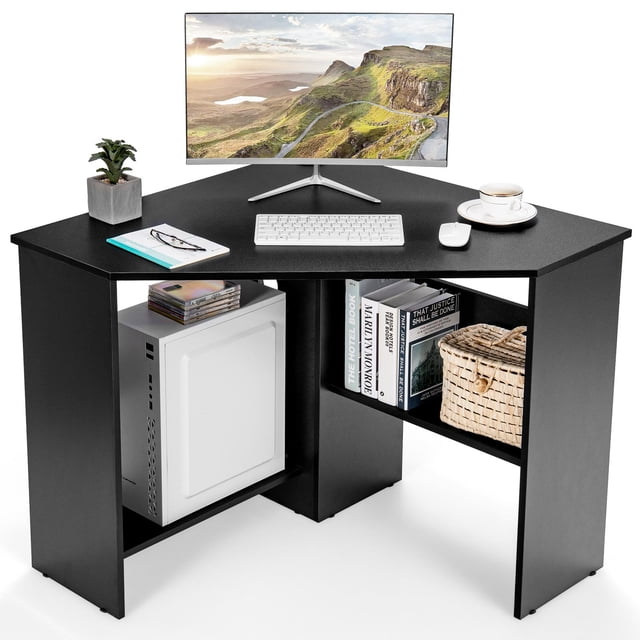 Ginatex Corner Computer Desk in black, featuring a space-saving triangular design with two storage shelves. The desk is shown in a home office setting with a monitor, keyboard, mouse, and decorative items on the tabletop. The lower shelves hold books, a computer host, and a woven basket. Made of engineered wood with non-slip foot pads for stability, this desk is ideal for small spaces.