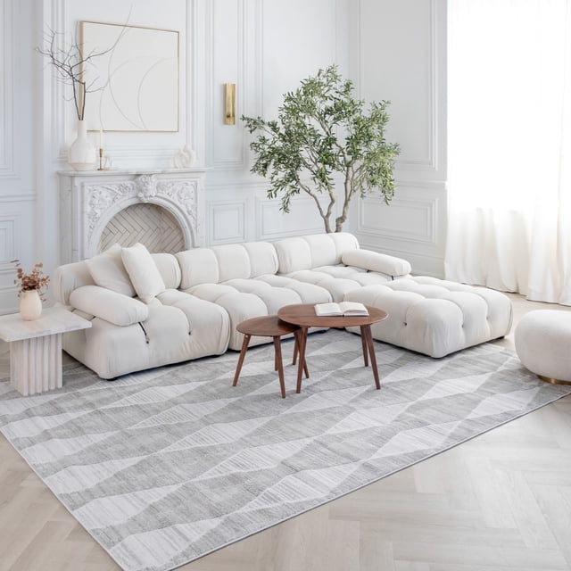 Rug Branch Astra Modern Indoor Area Rug in grey with an abstract design, placed in a bright living room with a tufted cream sectional sofa, two round wooden nesting tables, a white marble side table with dried flowers, a large green indoor plant, and white paneled walls featuring a decorative fireplace and sheer curtains.
