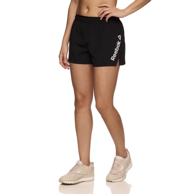 Reebok Women's Staple Running Short with Mesh Lining in black, made from lightweight 100% polyester fabric with elastic waistband and inside drawcord, side slit, and white Reebok Delta logo on the left leg, worn by a model with beige running shoes on a white background, ideal for running, training, or casual wear.