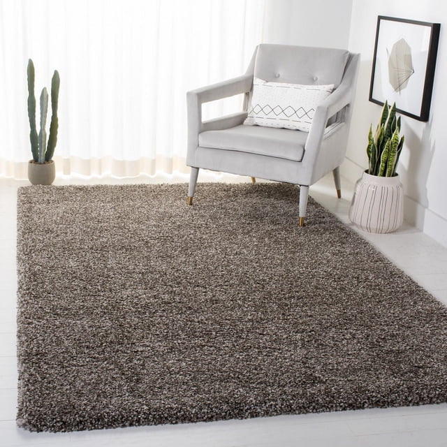 Safavieh Milan Harlow Solid Shag Area Rug in grey, 3' x 5', with a 2-inch thick dense shag pile made of durable synthetic yarns. Displayed in a bright room under a modern light grey armchair with gold-capped legs and a patterned lumbar pillow. Flanked by potted cactus and snake plants beside framed geometric wall art. Suitable for adding warmth and soft texture to contemporary home décor.