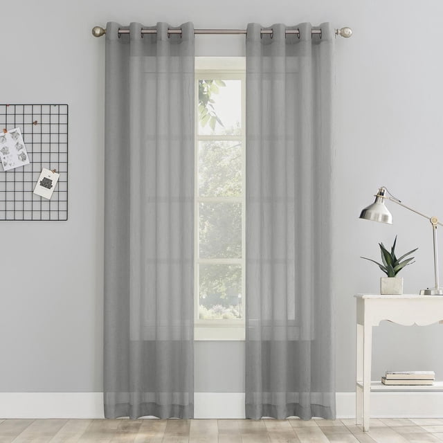 Mainstays Crushed Voile Window Curtains Panel Pair in light grey sheer voile fabric with metal grommets hanging on a curtain rod above a window, softly filtering light while enhancing privacy in a minimalist room with grey walls, white baseboard, a white side table with a silver desk lamp and potted plant, and a black wire grid with clipped notes on the left wall.