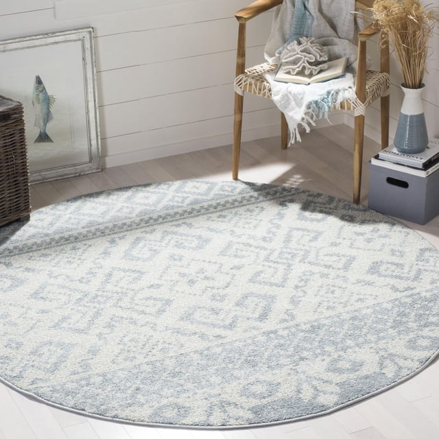 Safavieh Adirondack Zachary Southwestern Area Rug, round with a light grey and cream southwestern geometric pattern, power loomed polypropylene yarns, placed on light hardwood flooring beside a woven chair with a throw and coral decor, near books and a vase with dried grasses in a sunlit room, ideal for adding rustic lodge style to homes.