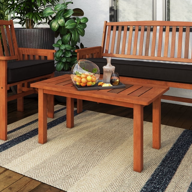 CorLiving Miramar Solid Wood Outdoor Coffee Table in brown with a slatted top designed to prevent water pooling, placed on a beige and navy striped outdoor rug. The hardwood table holds a tray with a glass bowl of oranges, a ceramic carafe, and a glass of dark beverage. It is positioned between matching wooden outdoor chairs and a bench with black cushions on a wooden deck with green plants nearby, ideal for small patios and decks.
