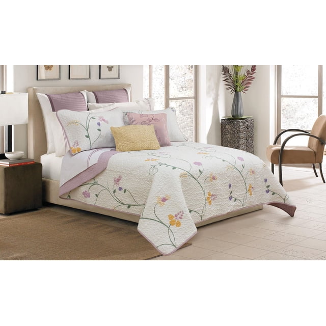 Safdie & Co. Home Deluxe Collection Multi Color 100% Polyester Quilt Set on a beige twin bed with a white quilted coverlet featuring embroidered yellow, purple, and pink floral patterns and mauve trim. Includes two mauve quilted pillow shams and decorative yellow and pink pillows. The room has a brown side table with books and lamp, a dark textured pedestal with a vase of greenery, and a beige armchair by large windows. This machine-washable set adds a soft floral accent to bedroom decor.