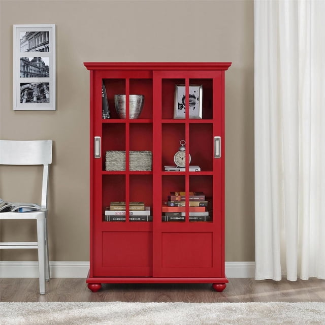 H-DOREL ACCENT CABINETS red wooden bookcase with sliding glass doors featuring square pane design, standing on rounded feet. It holds books, a silver bowl, framed photo, and clock, suitable for office furniture or bookshelf use in a neutral room with light wood flooring, white chair, and curtains.