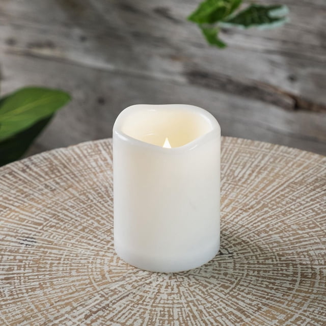 Hometrends LED candle flameless decorative candle with a realistic flickering flame inside a white plastic wax shell, featuring an on/off/timer switch, placed on a textured round wooden table with natural finish and blurred green leaves in the background.
