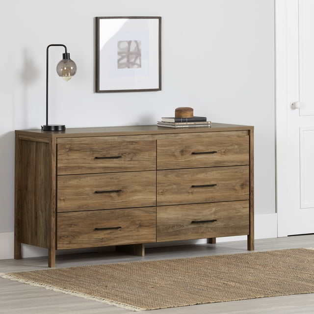 South Shore Gravity 6-Drawer Dresser for Adult in natural mango finish with six wide drawers featuring black slim handles and metal slides, supported by small squared legs. Styled with a modern black lamp, books, and a wooden decorative item on top, set against a white wall with framed abstract art, on a light wood floor with a woven beige rug. Ideal for organizing clothes, bedding, and accessories in a contemporary bedroom.