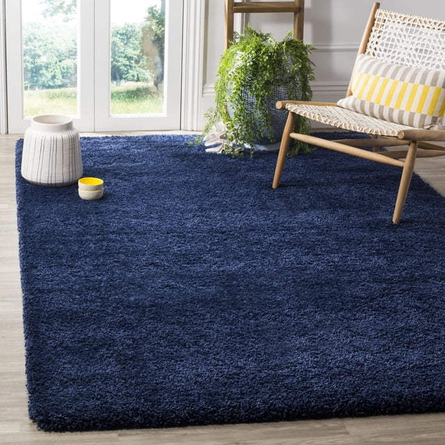 Safavieh Milan Harlow Tapis Shag Uni Bleu Marine Foncé avec texture de Fil Synthétique Peluche, Exposé sur Plancher de Bois Clair dans une Pièce Lumineuse avec une Chaise en Bois Tressée Présentant un Coussin Rayé Jaune et beige, une Grande Plante Verte dans un pot Bleu, et des vases Décoratifs en Céramique. Ce Tapis d'Accent Ajoute Chaleur et style à la Déco Contemporaine de la Maison et Convient aux Salons Ou aux Bureaux.
