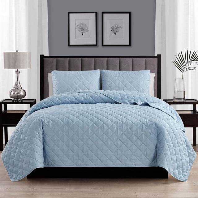 Swift Home Diamond Quilt Set in light blue with diamond-pattern quilting and matching pillow shams on a bed with a gray tufted headboard, accompanied by dark wood nightstands holding a silver lamp and a glass vase with a palm leaf, in a modern bedroom featuring framed botanical prints on a gray wall.